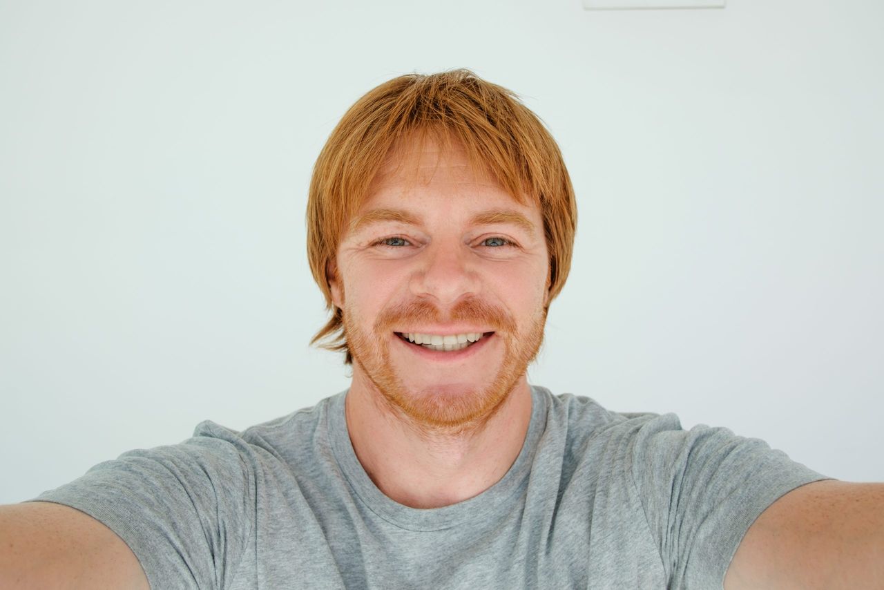 Closeup portrait of smiling middle-aged man looking at camera and taking selfie photo on gadget which is out of view. Isolated front view on grey background.