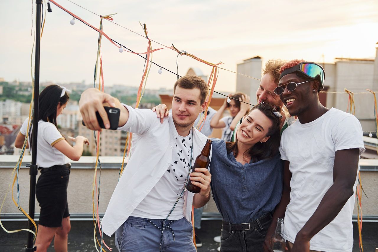 Group of young people in casual clothes have a party at rooftop together at daytime and doing selfie by phone.