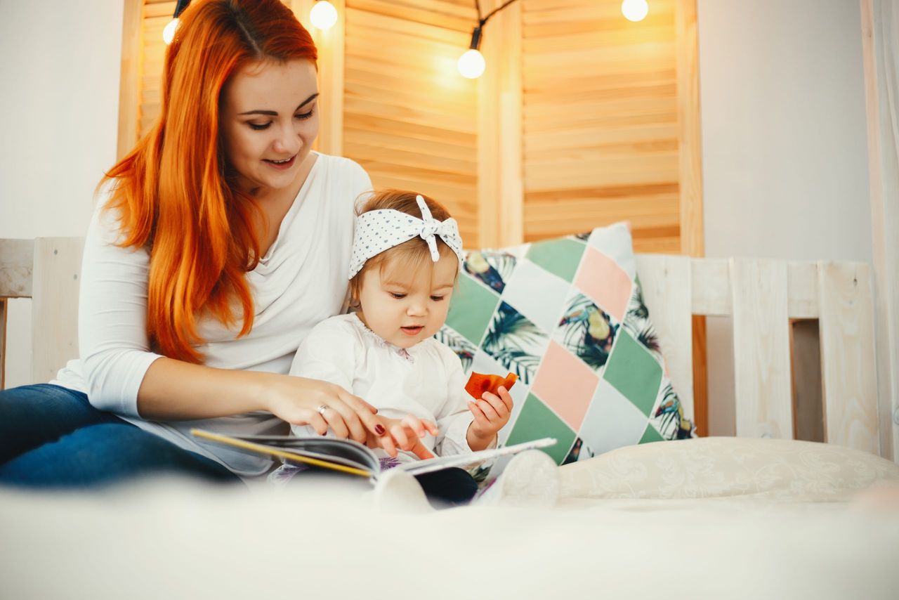 Beatuful ginger mother playing with daughter. Little girl sitting on a bed at home. Family reading the book