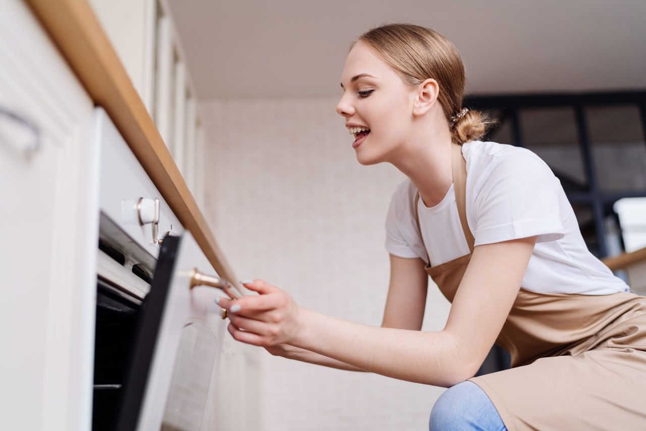 Young beautiful girl housewife in the kitchen in an apron baking happy cheerful emotions opening oven, look inside