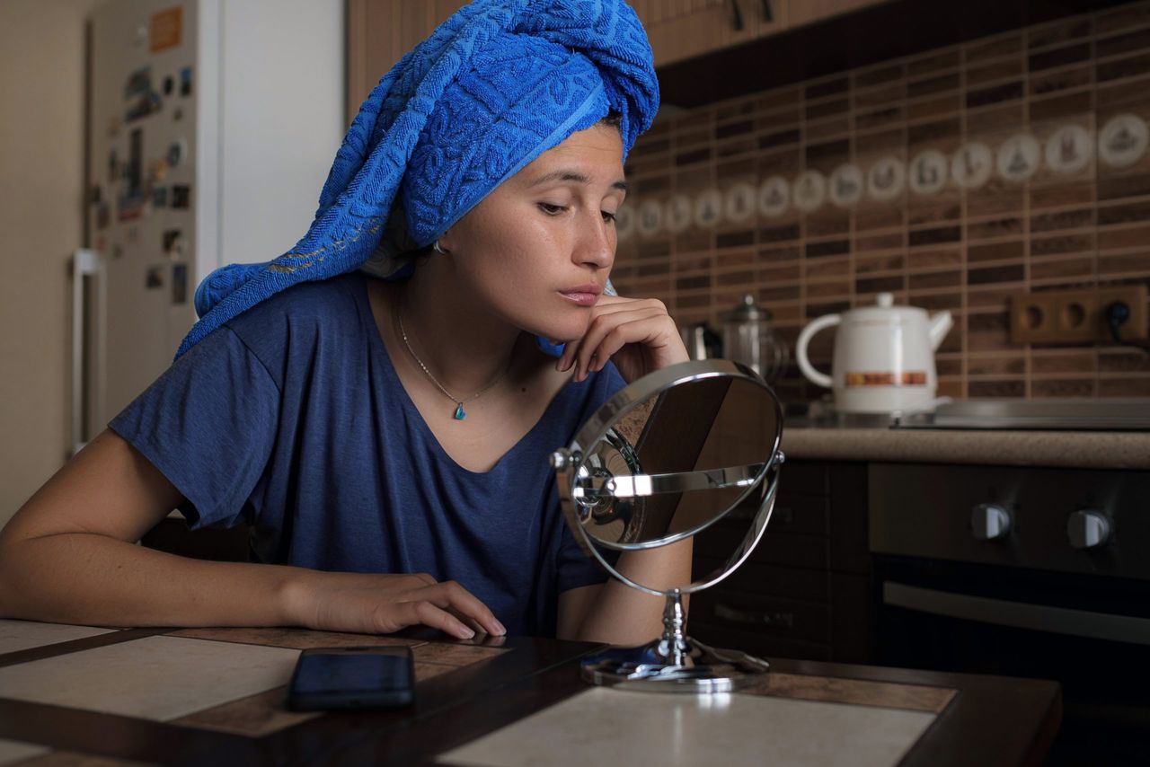 Beautiful asian woman wrapped in blue towel sitting on kitchen applying day cream and make face massage on her forehead, getting ready, morning routine.