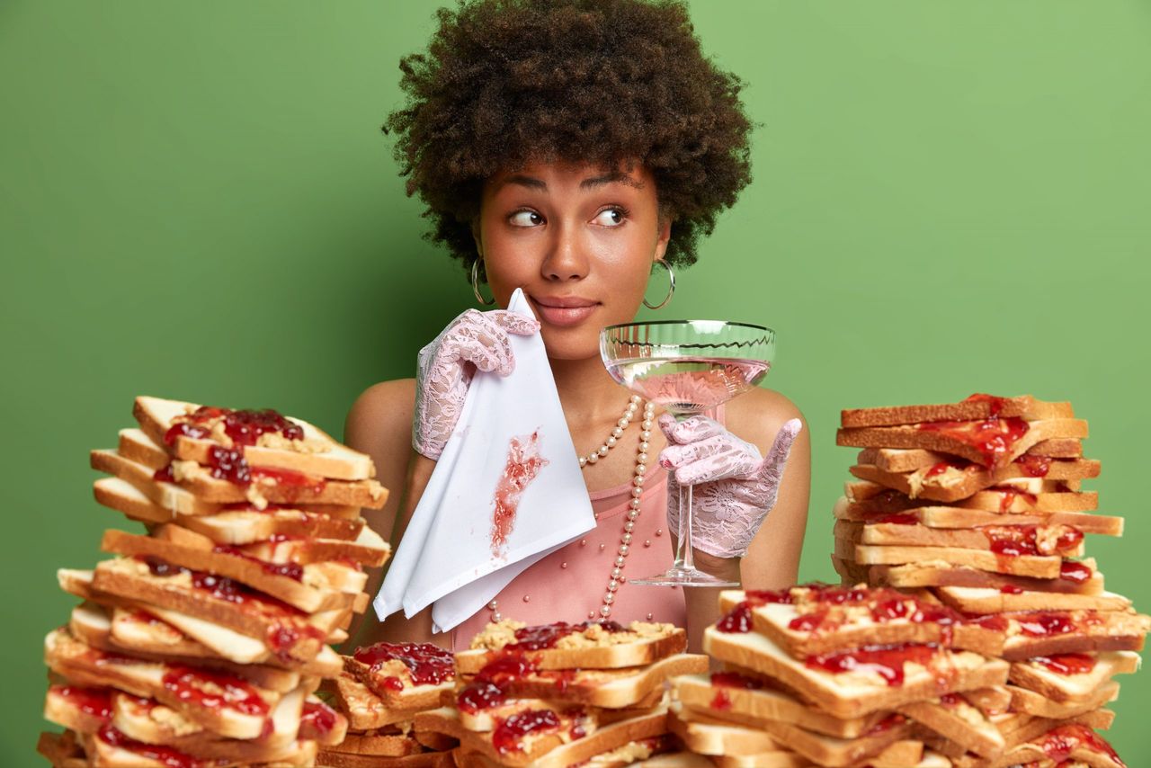 Elegant lady with Afro hairstyle, holds glass of martini and dirty napkin, comes in restaurant for informal meeting, surrounded by bread snacks, looks calm aside, isolated on green studio wall