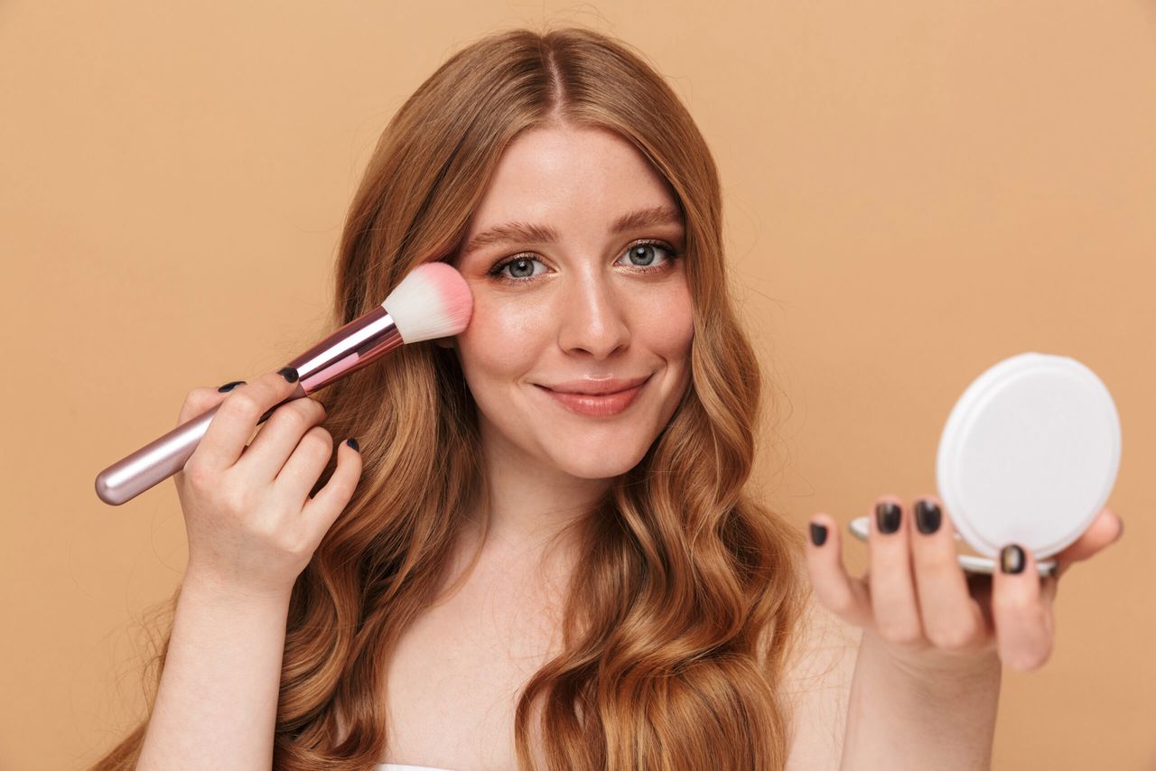 Portrait closeup of gorgeous young half-naked woman with long hair applying makeup with face brush isolated over beige background