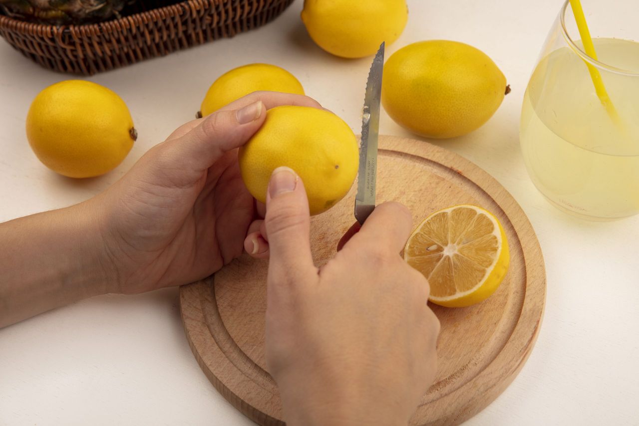 top view of female hands cutting fresh lemon on a wooden kitchen board with knife with lemons and lemon juice on a white background