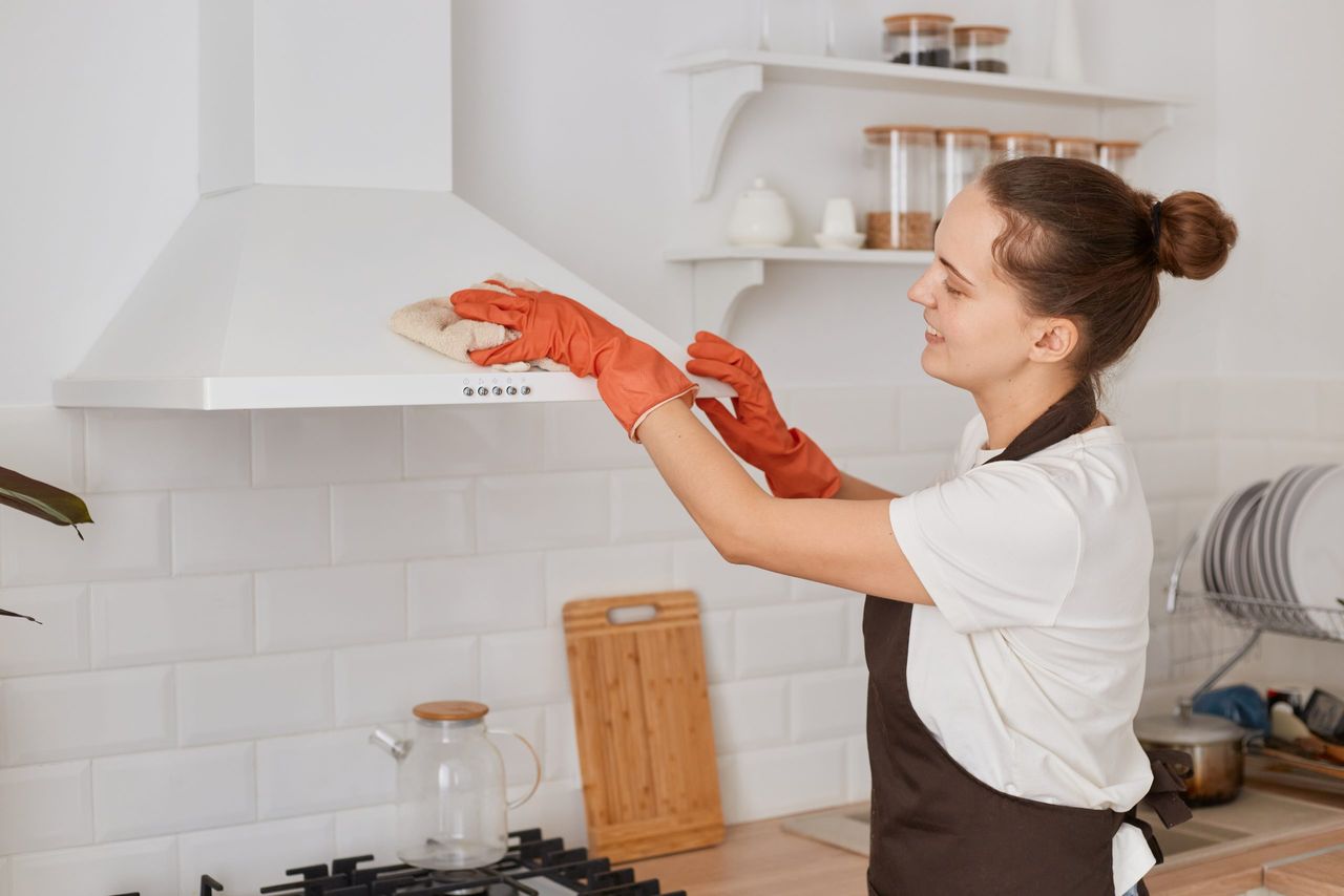 Horizontal shot of woman washing with a rag an electric cleaning hood at home kitchen, wearing white t shirt, brown apron and orange gloves, housekeeper with bun hairstyle doing her work.