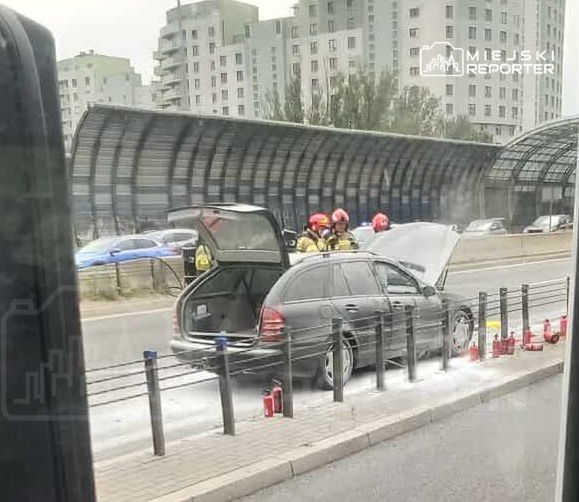 Czterech strażaków sprawdza samochód z otwartą maską na poboczu ulicy, obok widoczne gaśnice. W tle nowoczesne budynki.