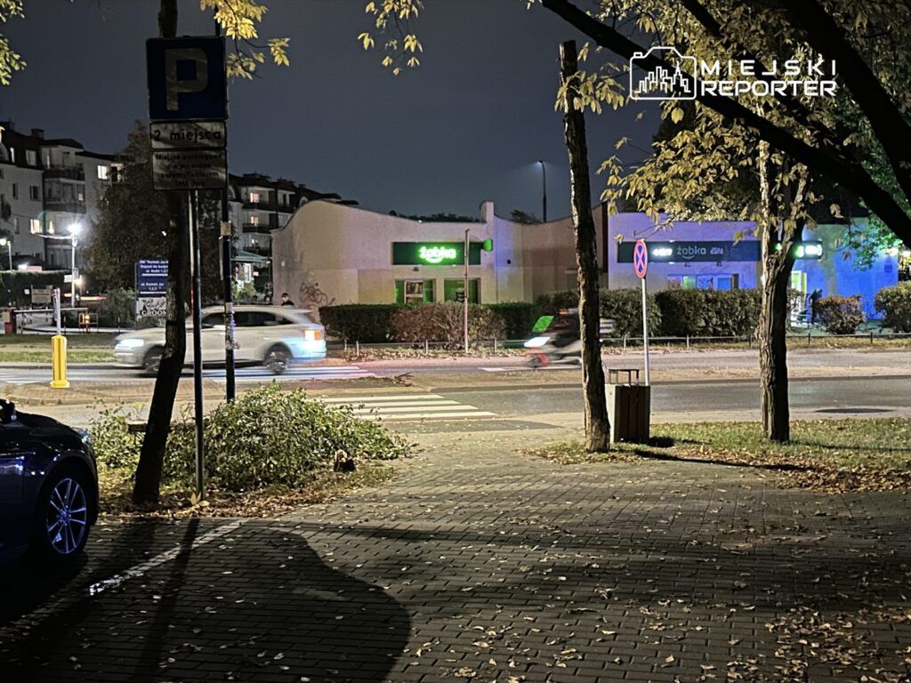 Zielony sklep z napisem "zabka" oświetlony nocą, w tle przejeżdżający samochód i znak parkingowy.