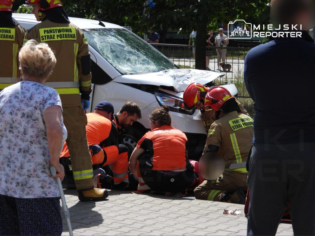 Grupa ratowników w mundurach pracuje nad osobą poszkodowaną w wypadku drogowym obok uszkodzonego samochodu.
