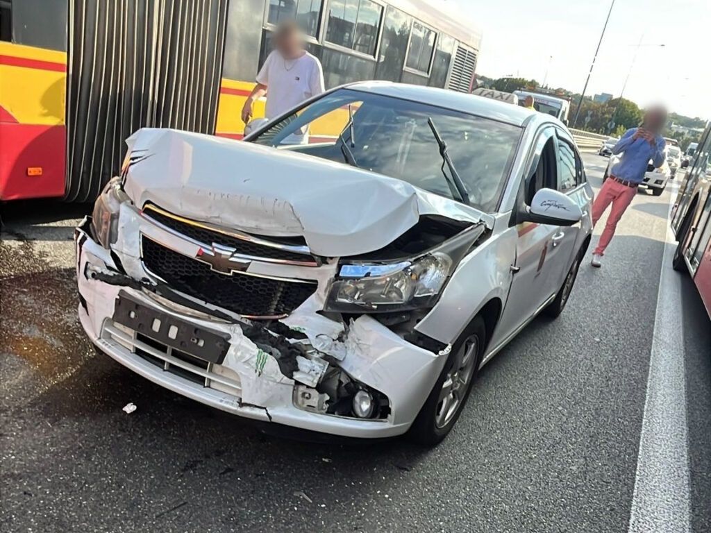 W wyniku kolizji drogowej uszkodzona została biała osobówka marki Chevrolet. Na zdjęciu widoczny jest poważnie wgnieciony przód pojazdu. W tle znajdują się autobusy komunikacji miejskiej, a w pobliżu kręci się kilka osób. Zdarzenie miało miejsce na ruchliwej ulicy.