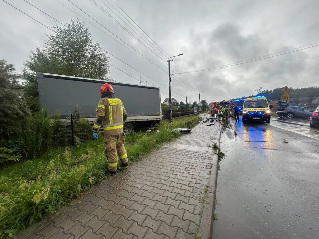 Strażak w mundurze z hełmem przemieszcza się wzdłuż drogi obok uszkodzonej ciężarówki, w tle widać ambulans i pojazdy ratunkowe.