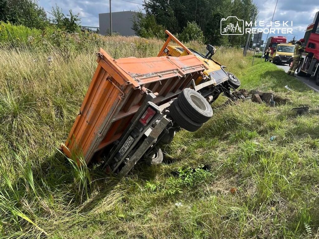 Złomowany pomarańczowy samochód ciężarowy leży na boku w wysokiej trawie przy drodze, obok stoją inne pojazdy.