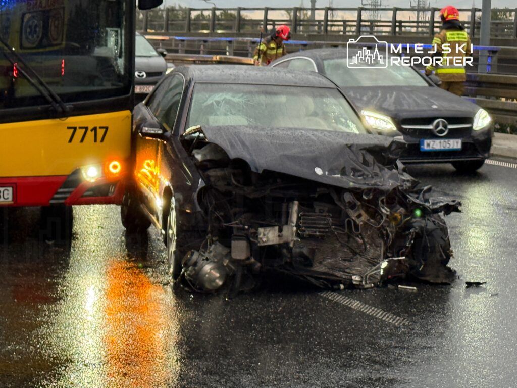 Czarny samochód osobowy z uszkodzoną przednią częścią stoi na mokrej jezdni obok autobusu, a strażacy przystępują do akcji.