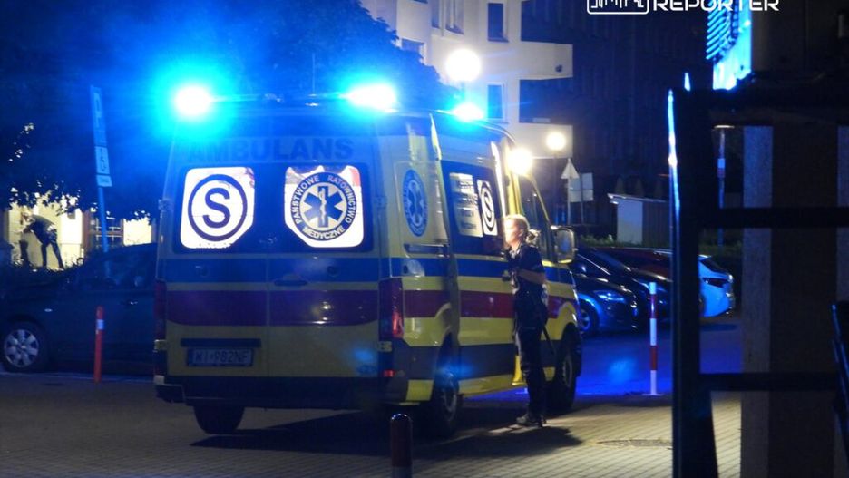 Karetka pogotowia z włączonymi sygnałami świetlnymi stoi na ulicy, a ratownik rozmawia z inną osobą w nocy.