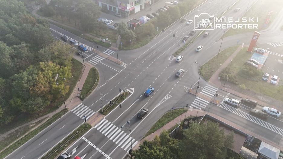 Ruch samochodowy na skrzyżowaniu z sygnalizacją świetlną, w tle nowoczesne budynki i drzewa.