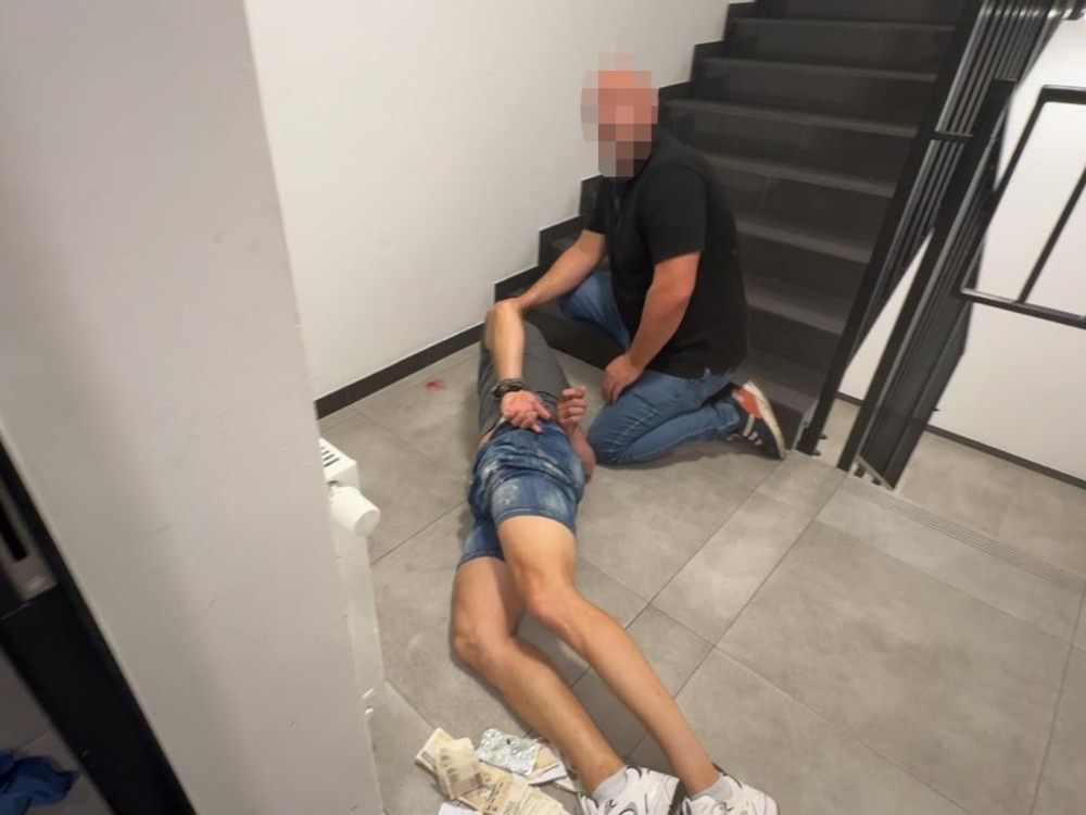 Mężczyzna w ciemnej koszulce pomaga leżącemu na schodach mężczyźnie w szortach, otoczeni przez szare ściany.