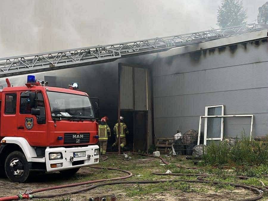 Czerwony wóz strażacki z niebieskimi światłami stoi przed budynkiem, z którego wydobywa się dym. Strażacy w mundurach przystępują do akcji gaśniczej.