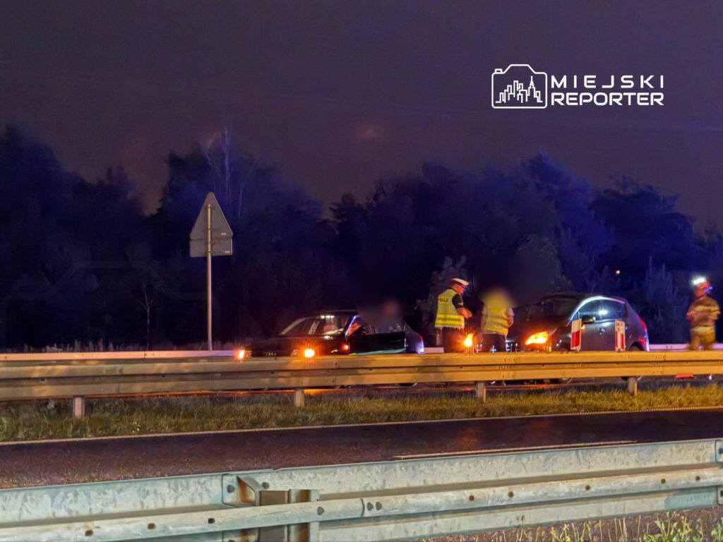 Na drodze nocą, funkcjonariusze policji i straży pożarnej prowadzą działania przy dwóch uszkodzonych samochodach.