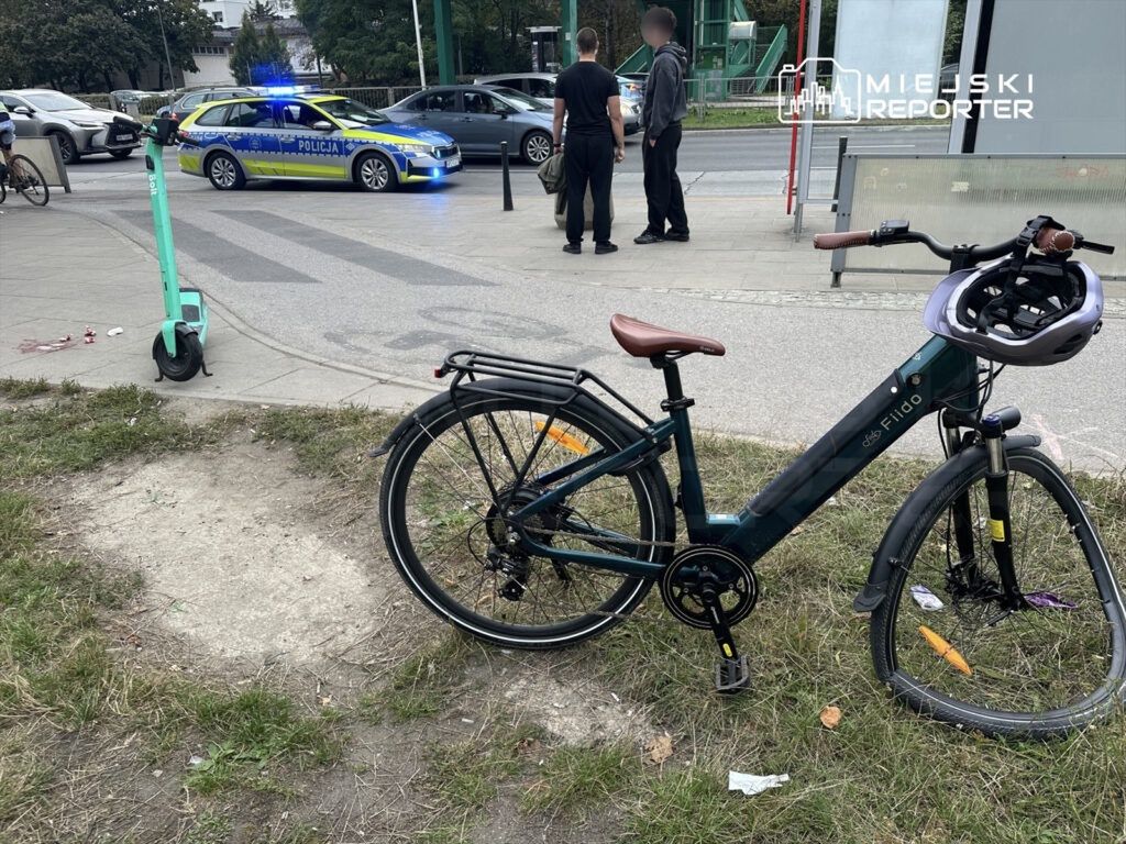 Policyjny radiowóz z niebieskimi światłami zatrzymany obok roweru na trawniku, w tle przechodnie i hulajnoga elektryczna.