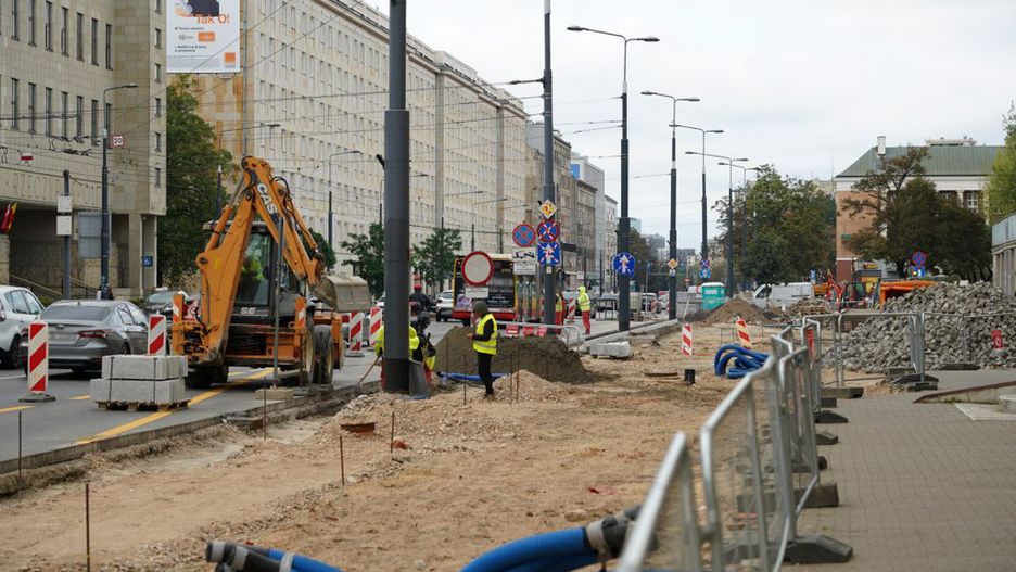 Ekipa budowlana pracuje przy wykopie drogowym, obok koparki i bloków betonowych, w miejskim otoczeniu.