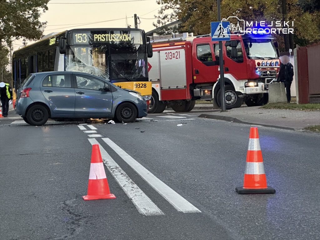 Zderzenie autobusu z osobowym samochodem na skrzyżowaniu, w tle widoczny wóz strażacki oraz ustawione pachołki.