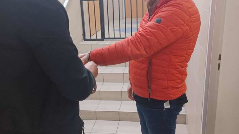 Mężczyzna w czerwonej kurtce stoi na schodach, podczas gdy inny mężczyzna zakłada mu kajdanki w budynku z kratami.