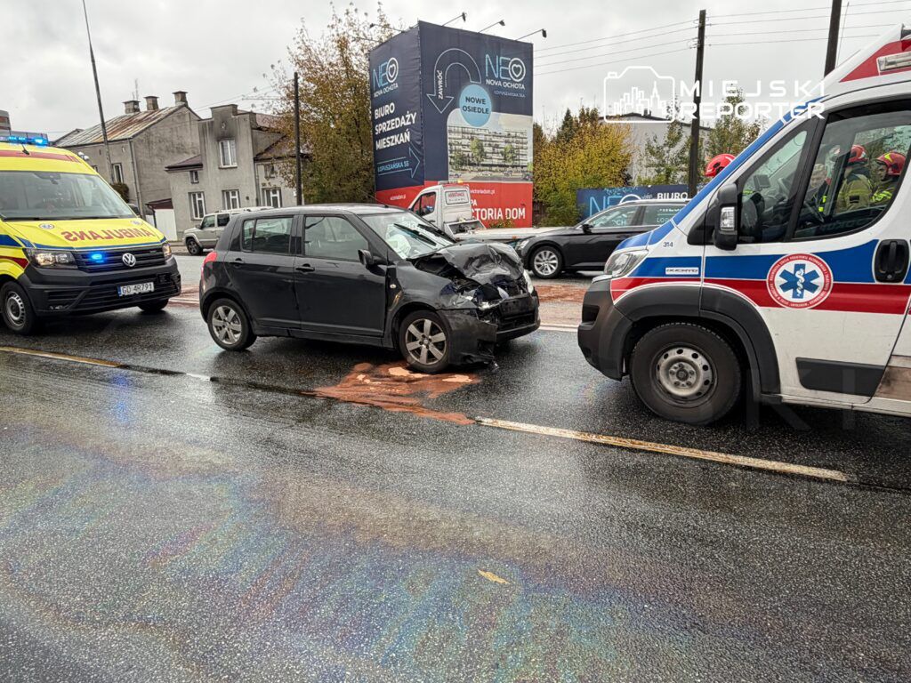 Wypadek drogowy z uszkodzonym czarnym samochodem osobowym oraz dwoma ambulansami na mokrej nawierzchni ulicy.