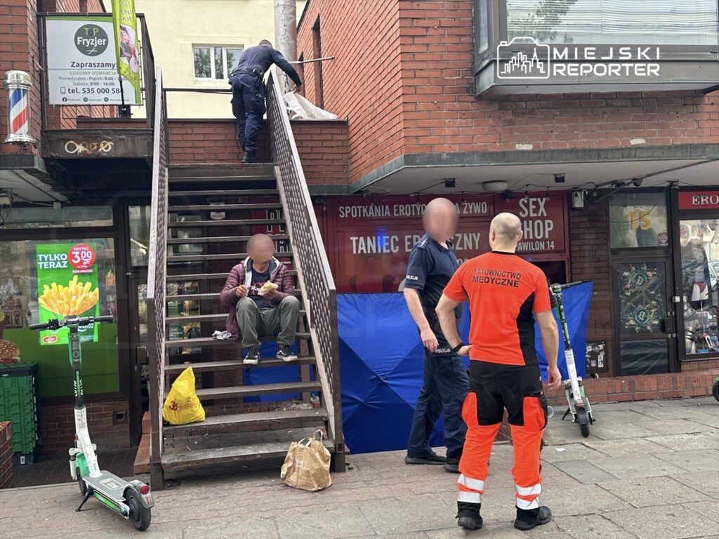 Mężczyzna siedzący na schodach, jedzący jedzenie, podczas gdy policjant i ratownik medyczny rozmawiają obok.