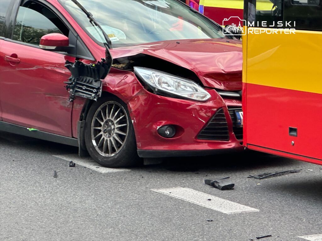 Czerwony samochód osobowy z uszkodzoną przednią częścią zderza się z autobusem na ulicy, obok leżą fragmenty plastiku.