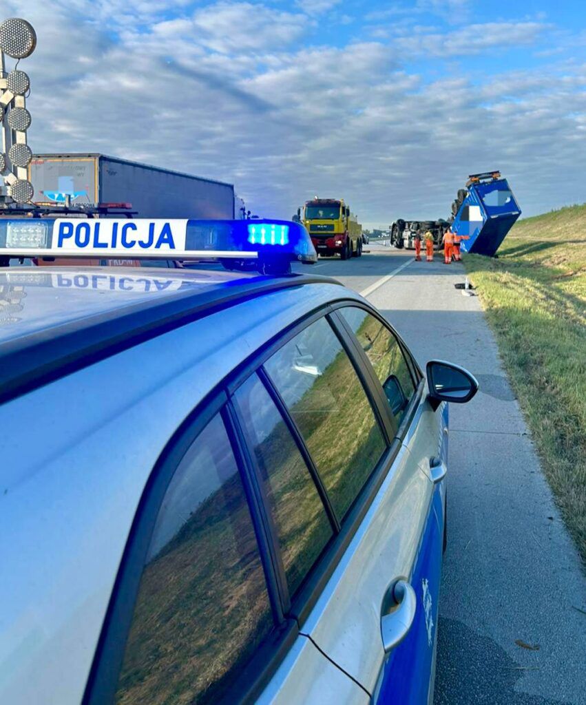 Na drodze stoi policyjny samochód z włączonymi sygnałami świetlnymi, obok pracują służby porządkowe przy przewróconym pojeździe.