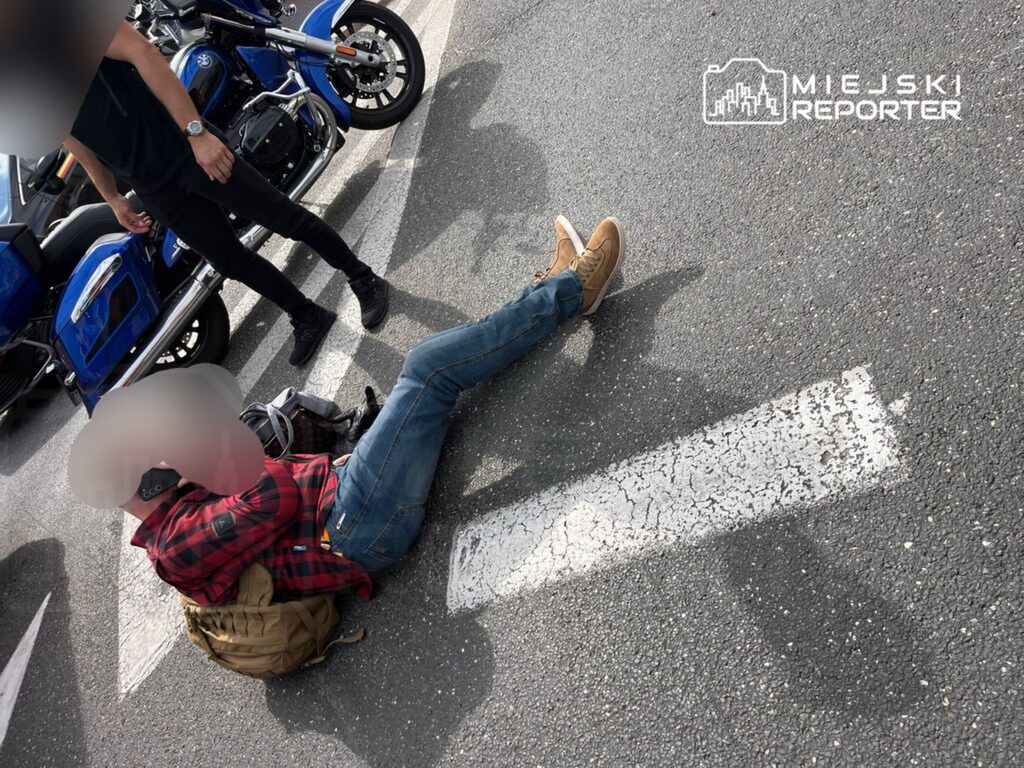 Mężczyzna leży na jezdni w pobliżu motocykli, obok stoi inna osoba. Na zdjęciu widoczne są elementy ruchu drogowego. Sytuacja sugeruje, że mogło dojść do incydentu. W tle widać pojazdy oraz fragment parkingu.