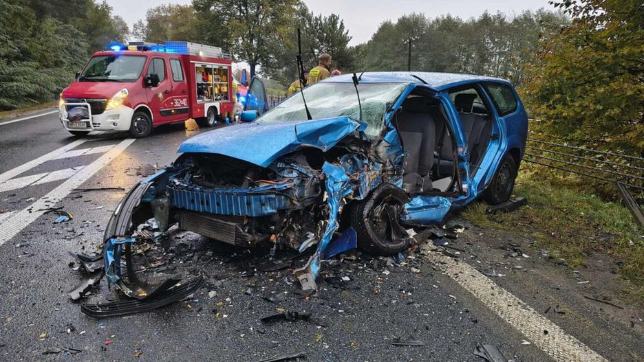 Zniszczony niebieski samochód osobowy leży na poboczu drogi, obok pojazd straży pożarnej oraz ratowników.
