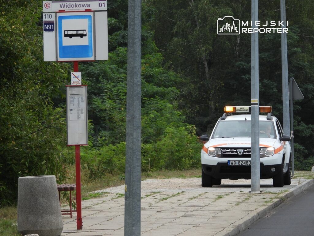Pojazd służbowy zaparkowany przy przystanku autobusowym w otoczeniu zieleni.
