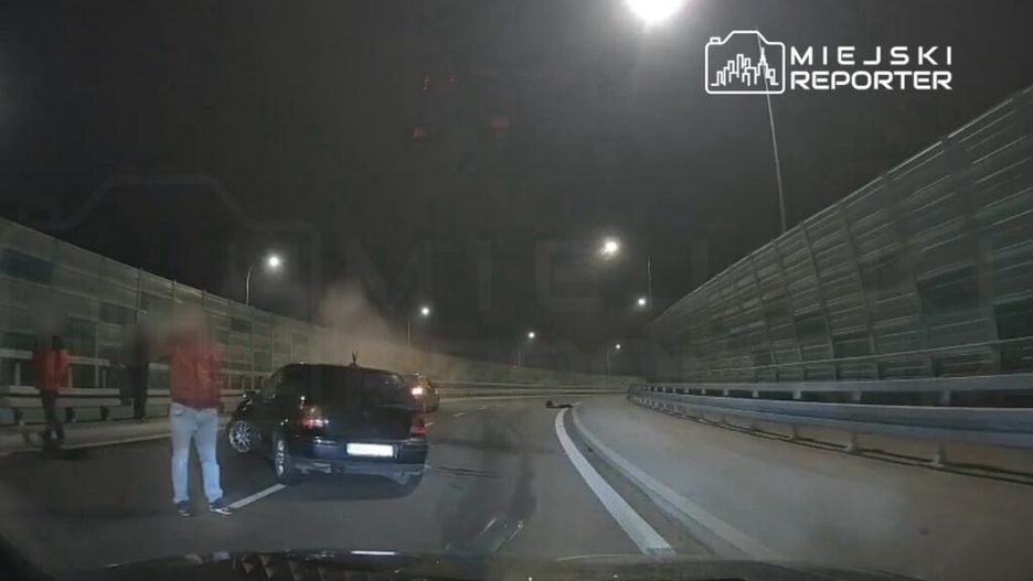 Grupa osób w czerwonych kurtkach stoi obok uszkodzonego samochodu na drodze, w tle widać dym.