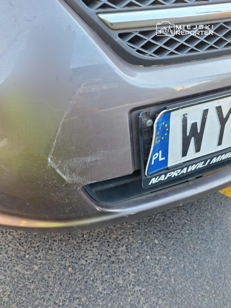 Zarysowany zderzak samochodu w kolorze szarym z widocznymi uszkodzeniami na dolnej krawędzi.