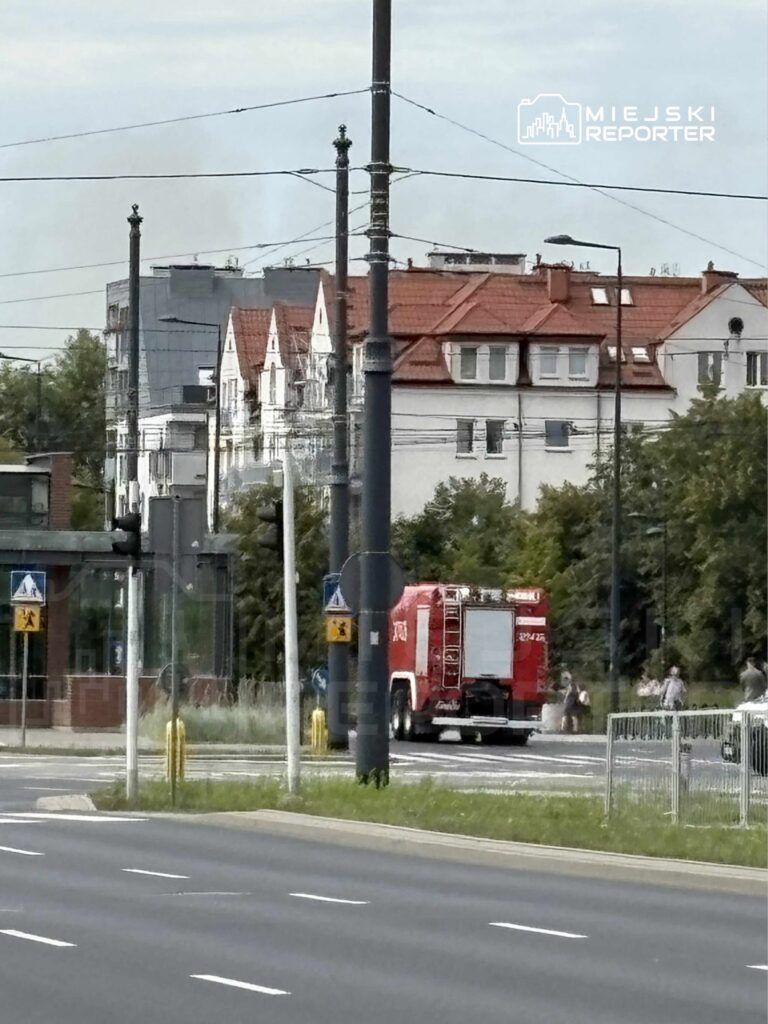 Czerwony wóz strażacki przejeżdża obok przystanku tramwajowego w miejskim otoczeniu.