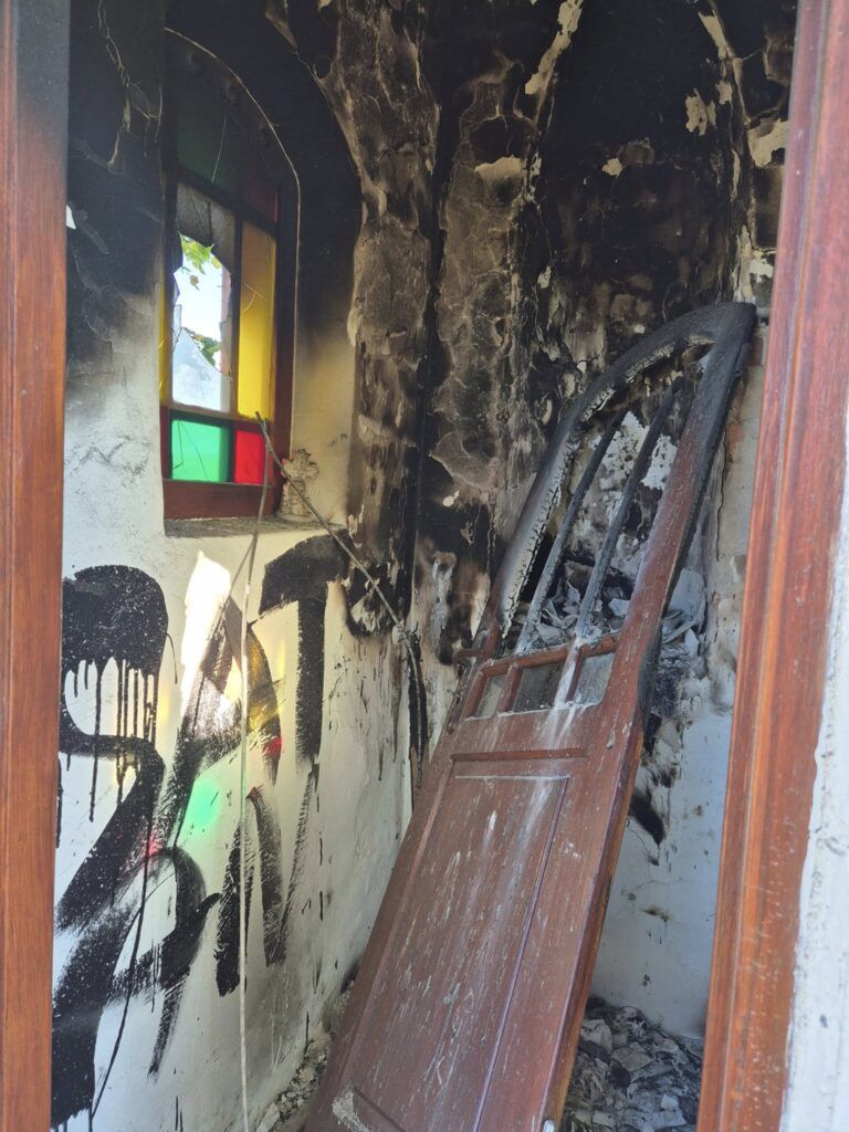 Zniszczona wnętrze budynku z poparzoną ścianą, przewróconymi drzwiami i oknem z kolorowym szkłem. Na ścianie widoczny napis.