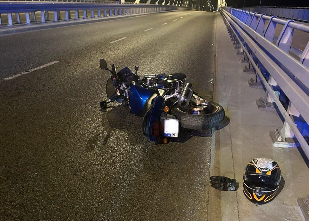 Motocykl leży na jezdni na moście, obok znajduje się kask i rękawica. Nocna sceneria z oświetleniem.