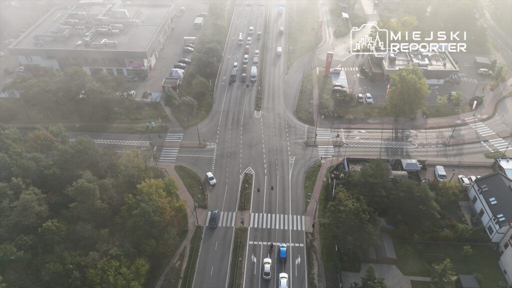 Widok z lotu ptaka na skrzyżowanie z ruchem samochodowym, otoczone drzewami i budynkami.