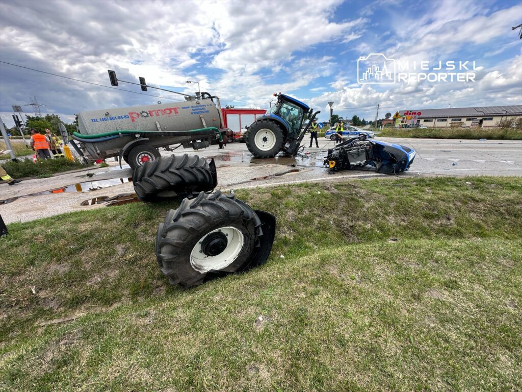 Traktor przewrócony na drodze, obok rozbite auto; w tle strażacy oraz pojazd gaśniczy.