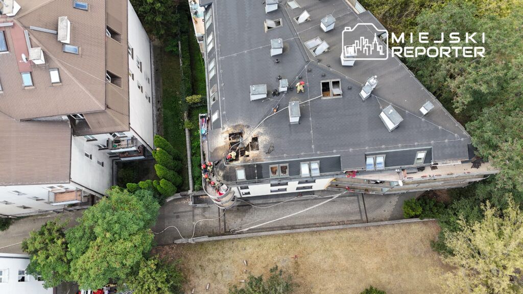 Grupa ratowników w kaskach pracuje na dachu budynku, usuwając zniszczoną część pokrycia.