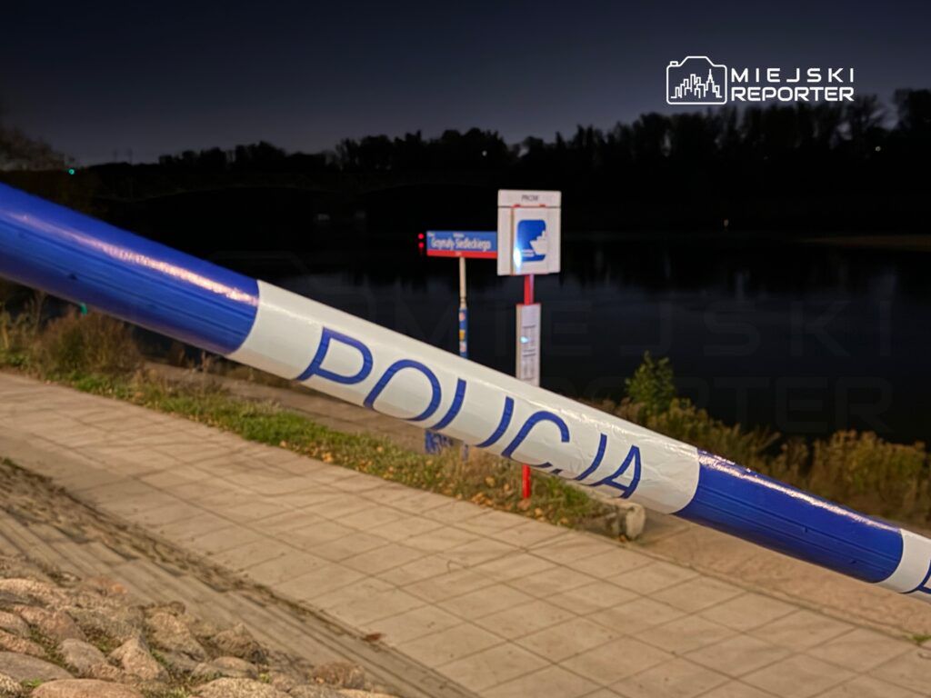 Policja zabezpiecza teren przy rzece, na tle słupa z informacją o przystanku. Nocna sceneria.