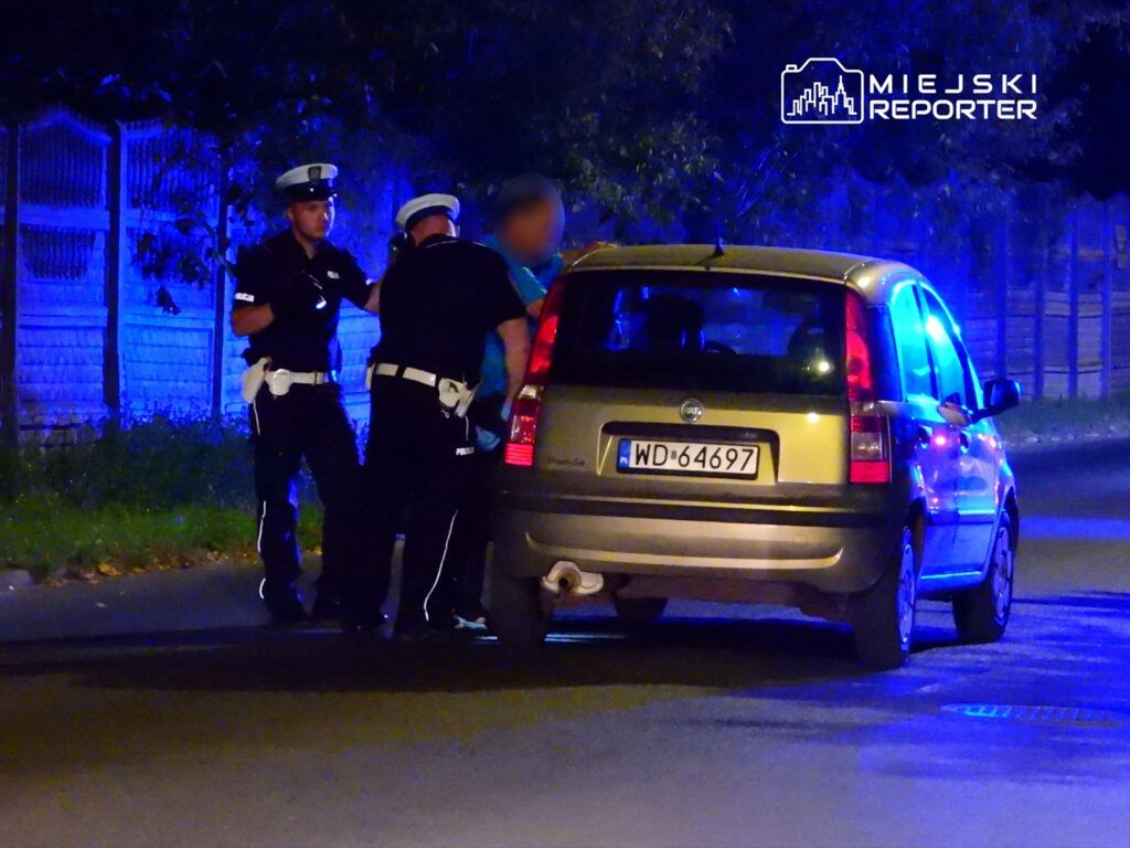 Dwaj policjanci kontrolują mężczyznę przy zaparkowanym samochodzie w nocy, w tle niebieskie światła.