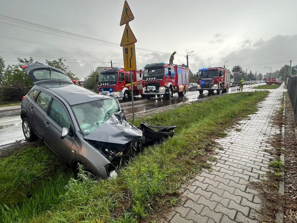 Uszkodzony samochód osobowy stoi na poboczu drogi, obok wozów strażackich na deszczowej ulicy.