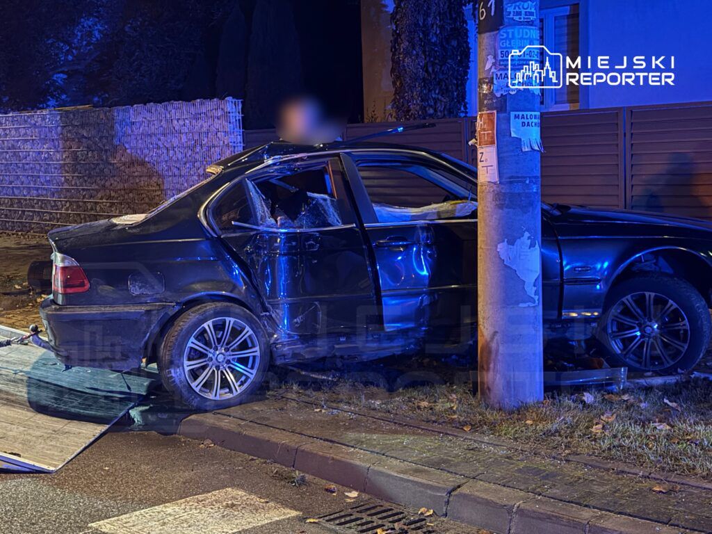 Czarny samochód osobowy uderzył w słup, z uszkodzoną tylną częścią i otwartymi drzwiami, na oświetlonej ulicy.