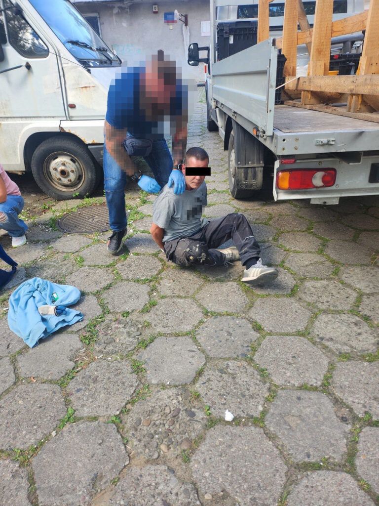 Mężczyzna w szarym T-shircie siedzi na ziemi z rękami związanymi, podczas gdy funkcjonariusz policji go kontroluje.