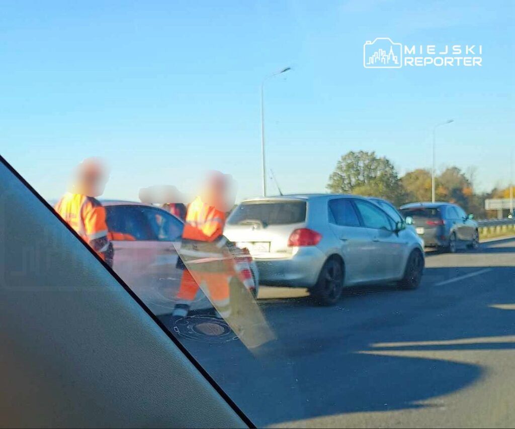 Dwaj mężczyźni w pomarańczowych kamizelkach odblaskowych kierują ruchem drogowym na zatłoczonej drodze.