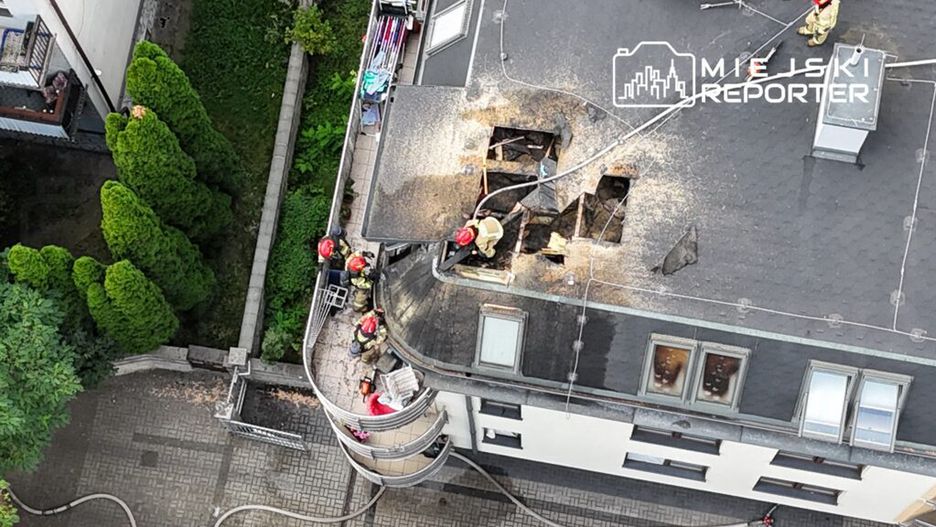 Grupa strażaków pracuje na dachu budynku, usuwając zniszczenia po pożarze, z widocznymi otworami w pokryciu dachowym.