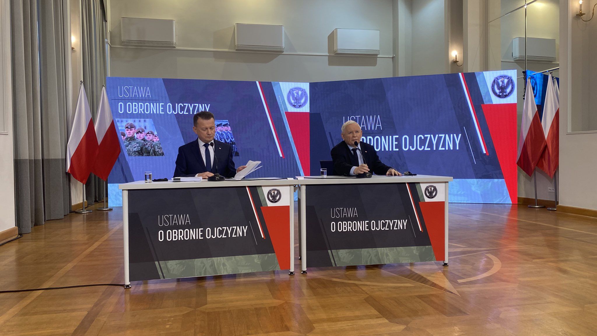 Na zdjęciu od lewej Mariusz Błaszczak i Jarosław Kaczyński. Politycy siedzą przy stole, ich nogi zasłania banda reklamowa MON z napisem "ustawa o obronie ojczyzny".