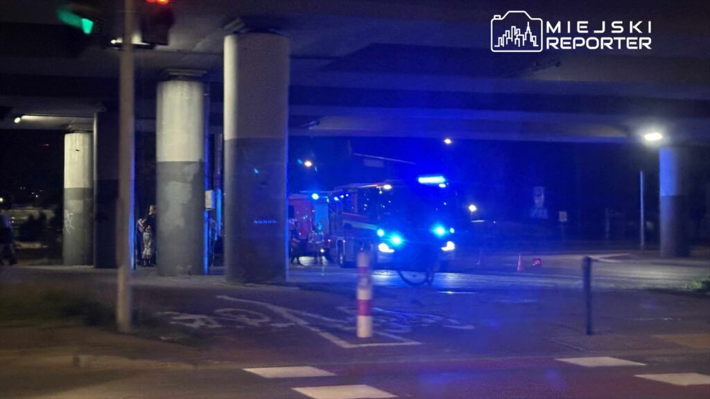 Straż pożarna z włączonymi sygnałami świetlnymi zatrzymana pod wiaduktem, obok grupy ludzi na chodniku.