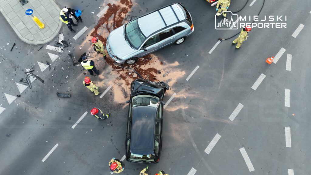 Na skrzyżowaniu dwóch ulic doszło do wypadku drogowego, w którym zderzyły się dwa samochody. Policjanci oraz strażacy pracują nad zabezpieczeniem miejsca zdarzenia. Wokół leżą fragmenty pojazdów oraz plamy płynów eksploatacyjnych.